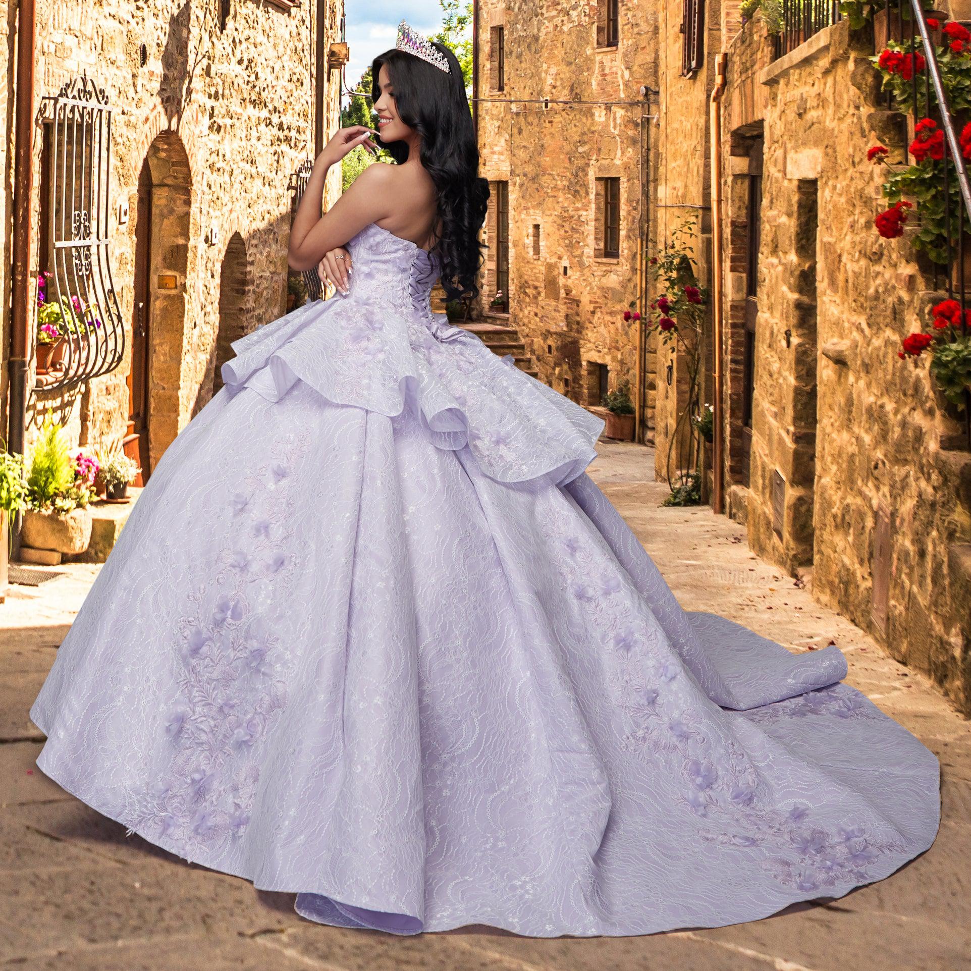 A woman in the Quinceañera Sample Dress QF115 and a tiara stands on a narrow cobblestone street lined with stone buildings and flowers.